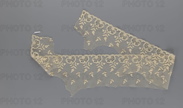 Fragment of lace, lace with plant ornaments, slightly serrated lower edge with shell motifs with sprouting twigs, above it projections of smaller twigs; 2nd half of the 19th century, Europe; lace, width 20 cm, length 161 cm, A piece of delicate lace fabric displayed against a smooth gray background; the lace is a pale cream color with an ornate scalloped border featuring repeating floral motifs and leaf-like shapes, and a sheer netted body scattered with small clustered floral elements; the fabric appears in a couple of irregular sections with frayed edges, showing both the decorative border and the translucent mesh between; overall colors visible are gray in the background and cream in the lace.