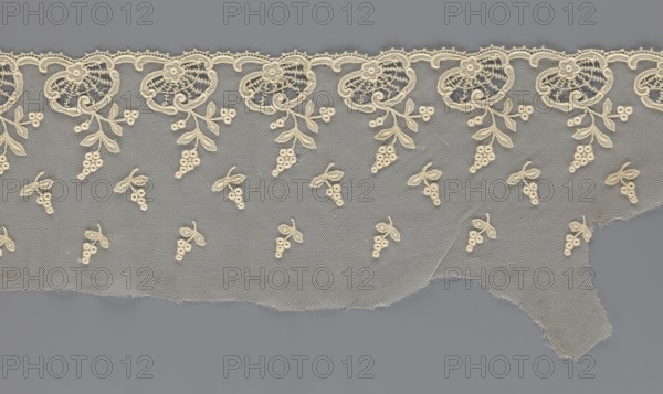 Fragment of lace, lace with plant ornaments, slightly serrated lower edge with shell motifs with sprouting twigs and above them projections of smaller twigs, 2nd half of the 19th century, Europe, width 20 cm, length 161 cm, A length of delicate lace in cream tones lies against a flat gray background; the lace features a scalloped upper edge with repeating ornate floral motifs and a mesh net ground sprinkled with a few small clustered berry-and-leaf designs, and the lower edge is uneven and slightly frayed.