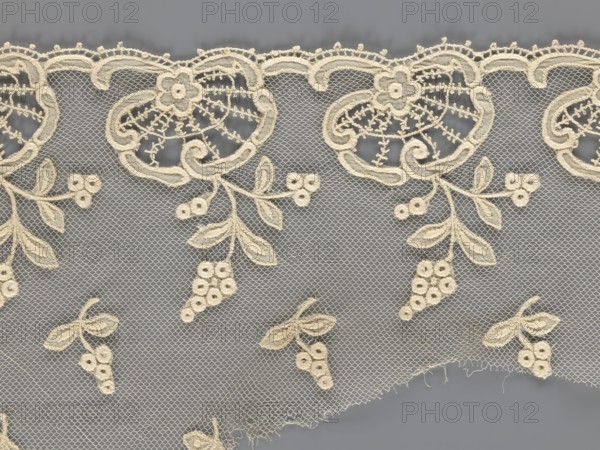 Fragment of lace, lace with plant ornaments, slightly serrated lower edge with shell motifs and sprouting twigs, above it projections of smaller twigs; 2nd half of the 19th century, Europe; lace, width 20 cm, length 161 cm, The image shows a piece of lace fabric with an intricate floral and vine pattern stitched in a cream color against a gray background. The lace features a scalloped top edge with embroidered flower motifs and looping ornamental shapes, and a net-like mesh fills the background. Several hanging clusters of embroidered berries and leaves extend downward, and a few small detached floral clusters are scattered across the lower portion. The lace has delicate openwork details and a slightly frayed lower edge.