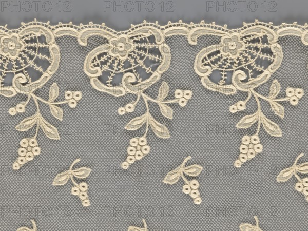Fragment of lace, lace with plant ornaments, slightly serrated lower edge with shell motifs with sprouting twigs and projections of smaller twigs, 2nd half of the 19th century, Europe, width 20 cm, length 161 cm, A delicate beige lace fabric with an allover mesh background and a scalloped edge at the top, featuring repeating embroidered floral and leaf motifs that hang down like sprigs with small clustered round shapes; the lace shows a contrast between the beige embroidery and a muted gray background, with the beige appearing slightly warm and the gray appearing cool.