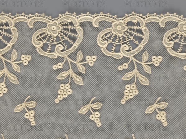 Fragment of lace, lace with plant ornaments, slightly serrated lower edge, featuring shell motifs with sprouting twigs and projections of smaller twigs above; 2nd half of the 19th century, Europe; lace, width 20 cm, length 161 cm, A section of delicate lace fabric with an intricate embroidered floral and vine pattern in a cream color against a muted gray background; the lace features a scalloped top edge with ornate looped motifs and small flower shapes, a netted mesh filling, and hanging vine elements with stylized leaves and clusters of round embroidered berry-like shapes, all in cream tones contrasted with the gray behind.