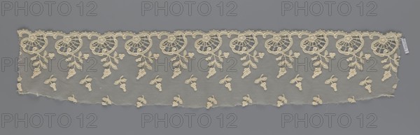 Fragment of lace, lace of, plant ornaments, slightly serrated lower edge with shell motifs and sprouting twigs, above it projections of smaller twigs. 2nd half of the 19th century, Europe. Lace, width 17 cm, length 86 cm., A long strip of delicate lace in cream against a gray background, featuring a scalloped top edge with a repeating ornate floral motif and trailing leaves, and a mesh lower field scattered with a few small cluster-and-leaf designs; the lace shows frayed edges at the bottom and sides and fine embroidery details in the cream thread.