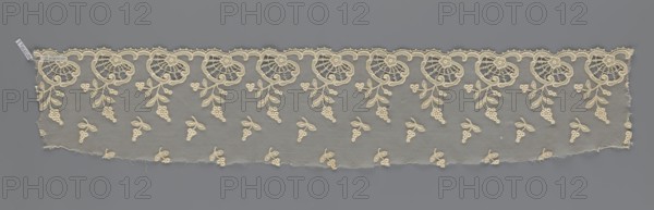 Fragment of lace, lace of, plant ornaments, slightly serrated lower edge with shell motifs and sprouting twigs, above it projections of smaller twigs. 2nd half of the 19th century, Europe. Lace, width 17 cm, length 86 cm., A long horizontal piece of lace laid flat against a solid background, showing a scalloped upper edge with an ornate repeating floral motif and a lower straight edge with small fragments and a few loose threads; the lace fabric is a delicate net with an all-over pattern of small clustered berry-like shapes and paired leaf motifs scattered across the field, while the top border features larger rounded floral designs with radiating internal stitching and trailing leaf sprays; visible colors include cream in the lace and a mid-tone gray for the background.