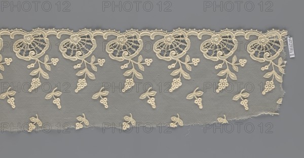 Fragment of lace, creator unknown, lace of, plant ornaments, slightly serrated lower edge with shell motifs and sprouting twigs, above it projections of smaller twigs; 2nd half of the 19th century, Europe; lace, width 17 cm, length 86 cm., A strip of delicate lace with an open net background in gray and cream tones, featuring a repeating floral border along the top with ornate fan-shaped blossoms and scalloped edging in cream, and a pattern of small clustered berry motifs and paired leaves scattered across the lower area; the lace shows frayed edges and loose threads along the bottom and right sides, and the overall colors visible are cream and gray.