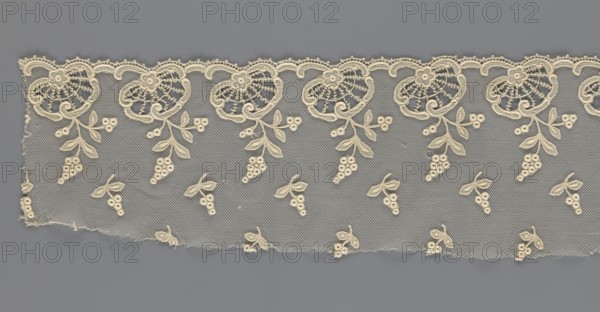 Fragment of lace, lace of, plant ornaments, slightly serrated lower edge with shell motifs and sprouting twigs, above it projections of smaller twigs; 2nd half of the 19th century, Europe; lace, width 17 cm, length 86 cm, A horizontal strip of delicate lace on a flat background shows a repeating floral and berry motif; the lace is a light cream color with fine netting and ornate scalloped edging along the top, including larger circular floral shapes with radiating stitch details and small flower centers, from which leafy sprigs and cluster-like berry shapes hang; the lower area of the lace contains scattered small leaf-and-berry clusters; the background behind the lace is a smooth gray, and the overall colors visible are cream and gray.