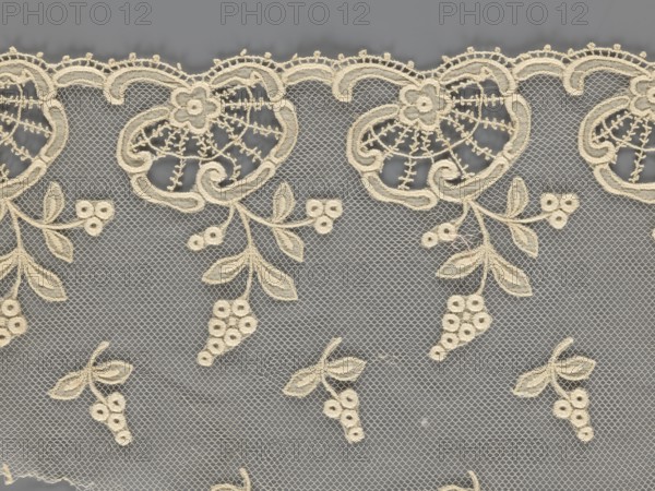 Fragment of lace, lace of, plant ornaments, slightly serrated lower edge with shell motifs and sprouting twigs, above it projections of smaller twigs. 2nd half of the 19th century, Europe. Lace, width 17 cm, length 86 cm., A delicate lace fabric with an off-white embroidered floral and vine pattern set against a gray background, featuring a scalloped top edge with ornate curving motifs and small embroidered flowers, trailing stems with leaves and clusters of round embroidered berry-like shapes, a fine hexagonal mesh filling the spaces between the motifs, and visible subtle variations in the off-white thread and gray behind it.