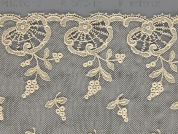 Fragment of lace, lace, plant ornaments, slightly serrated lower edge with shell motifs with sprouting twigs; above it projections of smaller twigs. 2nd half of the 19th century, Europe. Lace, width 17 cm, length 86 cm., A section of delicate lace fabric is shown with an off-white embroidered floral and grapevine pattern on a gray mesh background; the design features a scalloped top edge with ornate circular floral motifs, trailing stems with clustered round berry shapes and elongated leaves, and several isolated small grape cluster motifs scattered below, all in off-white against the gray.