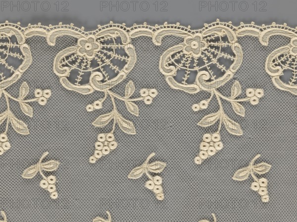 Fragment of lace, lace of, plant ornaments, slightly serrated lower edge with shell motifs with sprouting twigs; above it projections of smaller twigs. 2nd half of the 19th century, Europe. Lace; width 17 cm, length 86 cm., A patterned textile with a sheer gray background featuring an embroidered cream lace overlay; the lace shows a scalloped top edge with ornate curved floral motifs, branching stems with leaves and clusters of small round flower-like shapes, and several isolated small floral clusters scattered across the mesh.