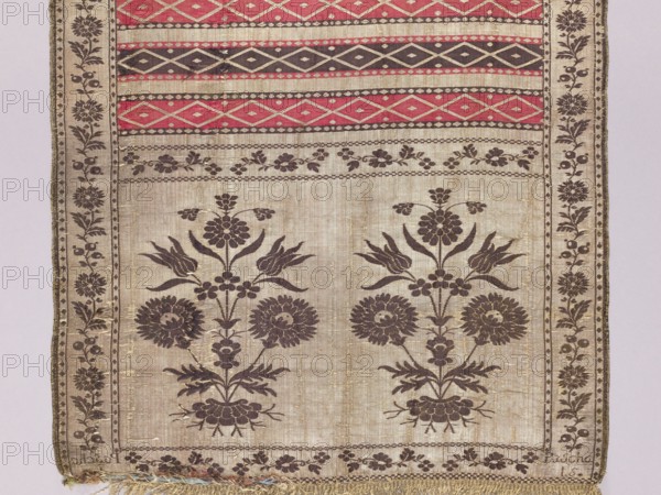 Kontusz sash, Paschalis Jakubowicz Manufactory Warsaw; belt manufacturer; 1788-1794. Solid kontusz sash, double-sided. Bipartite heads with a flower bush pattern. Alternating transverse stripes, plain and with geometric ornamentation. Edge with a floral tendril motif. Plant ornaments, geometric ornaments, flower bushes, floral ornaments. Colors: gold, silver, blue, yellow-green, light red, brown, light blue. Sew-on fringe, silver, with gaps. Ornament: - Heads: two motifs of rooted flower bushes. - Center: alternating transverse shelves, smooth and with rhombus motifs. - Borders: vertical and horizontal with different floral tendril motifs; checkerboard and rhombus. Colors placement: - Front: background of heads, borders and smooth shelves silver, ornament brown; background of the shelves with rhombuses alternating red and brown, silver ornament. - Reverse right: only smooth shelves alternating red and brown. Signed in the heads: Pascha/Nov. Weave density: approx. 19/34 x2. Date: 1788–1...