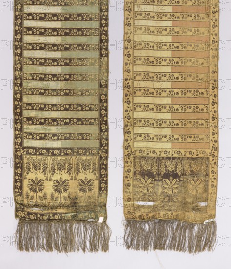 Kontusz belt, PC Salzhüber Manufactory Gdansk; belt manufacturer; ca. 1775–1800. Single heads with three tall stylized trees and cactus bushes between them; at the upper edge of the heads motifs of a suspended flower garland and cords with tassels. Alternating transverse stripes, plain and with floral tendrils; floral tendril edging. Gold fringe sewn on. Colors: straw, brown, cream, sky blue, crimson-pink. Before 1793. Gdansk place of origin. Clothing accessory, national costume; length 343 cm, width 32.5 cm., Two long vertical textile panels are shown side by side against a pale background. Each panel has alternating horizontal bands with floral motifs and solid stripes, framed by narrow decorative borders. The left panel features a darker palette with muted green and deep brown bands and gold floral patterns, ending in a rectangular section with stylized floral and leafy designs above a fringe of long tassels in brownish-gray. The right panel displays lighter beige and tan tones with...