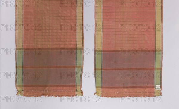 Kontusz belt, Polish/Ukraine Volhynia? origin, mid-18th century. Men's clothing accessory. Silk with smooth heads, half-cut by a 1 cm wide line; smooth stripes separated by narrow stripes; along both sides a green trail 3 cm wide. Length 240 cm, width 42.5 cm., A pair of long rectangular fabric panels hangs side by side against a pale background, each panel showing a horizontal striped pattern with a dominant warm reddish-orange field transitioning to a muted brownish band near the lower portion; both panels have vertical border stripes in pale yellow and green with thin dark lines, and the fabric appears semi-sheer with a subtle grid texture. The lower edges of the panels show frayed hems with loose threads and small gathered corners, and there is a small pale patch near one lower edge. Visible colors include reddish-orange, brown, pale yellow, green, dark thin accent lines, and the pale neutral background.