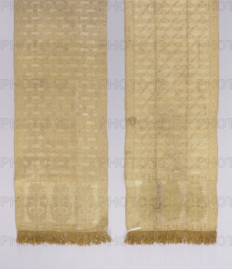 Kontusz belt. Silk with gilded silver thread; plain weave, brocaded single fabric; density 26 warps and 56 wefts per cm. 18th century. Length 314 cm, width 31 cm. Materials: silk, gilded silver thread; fringe sewn-on yellow silk with some gold threads. Description: solid, double-sided men's belt used as clothing/fashion accessory in Polish national costume. Ornament: three bush motifs each with tulip flowers as heads; center with transverse shelves alternating with leafy tendrils, palmettes, and projections of rosettes; border with a floral tendril motif. Patterns arranged in wavy snake or diagonal stripes. Colors: right side background white with gold ornament. Possible places of origin: Grodno, Italy, France, Grand Duchy of Lithuania, Poland., The image shows a pair of long, narrow woven textiles placed side by side against a light gray background; both pieces are predominantly pale gold and beige with subtle woven patterns creating vertical stripes and floral motifs, each ending in ...