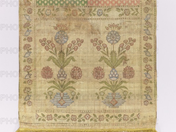 Kontusz sash, Bipartite heads with the pattern of flower bouquets on a vase-base, alternating transverse stripes with an ornament of floral tendrils and dots. Ribbon made of waxflower. Colors: brown, blue, red, green, and ochre. Signed at the bottom in the heads: FS lying. Late 18th century, Kraków. Polish sash kontusz sash for national costume; men's clothing accessory. Length 370 cm, width 35 cm. Ornaments: flower bouquets, flower bushes, szelagki, geometric and plant ornaments. Revindication, A textile panel with a faded beige background showing a symmetrical floral design, featuring a group of stylized flowers and leaves in muted pink, pale blue, and soft green tones, framed by a narrow floral border in similar colors and small blue bird-like motifs, and finished at the bottom with a short fringe in golden yellow.