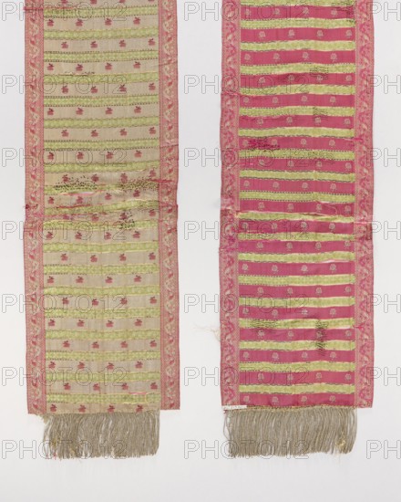 Kontusz belt, 18th century. Semi-solid, double-sided men's clothing accessory kontusz belt with floral ornaments and Vinca edging; gold fringe. Colors: red, green, and gold. Design: transverse stripes—green with rosettes and plain red with floral projections. Length 275 cm; width 26 cm. /place of origin., The image shows two long narrow woven fabric panels hanging vertically side by side against a light background. Each panel has a decorative border in pink with floral motifs and alternating horizontal bands of green and a pale beige or deep pink field with small repeating floral or geometric motifs. The left panel primarily features pale beige fields between green bands with pink borders, while the right panel primarily features deep pink fields between green bands with similar pink borders. Both panels show areas of wear and irregularities in the weave, with a few darker spots and slight fraying along seams. The bottom edges of both panels end in long silvery-grey fringe tassels. Vis...