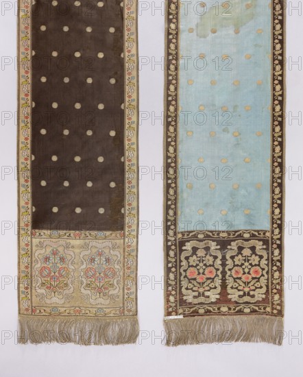 Kontusz belt;, ; ornament: flower bushes, geometric and plant ornaments; bilateral, semi-solid belt; Polish kontusz belt national costume clothing accessory; bipartite silver-gold heads with floral bouquets enclosed in a wavy line; bouquet colors crimson, blue, and green; one side background brown-gray-black with gold dots, other side blue with gold dots; signed in the heads: FS lying down; 18th century, Poland; length 291 cm, width 31 cm., Two long narrow textile panels hang side by side against a pale background; the left panel is primarily dark brown with a pattern of evenly spaced circular motifs and a narrow floral border in beige with touches of red, blue, and yellow, while the lower section of this panel features a beige rectangular area decorated with stylized floral medallions in muted red, blue, and green and a fringe of light beige threads at the bottom. The right panel is mainly pale blue with similar circular motifs and a brown floral border, showing a faded pale green are...