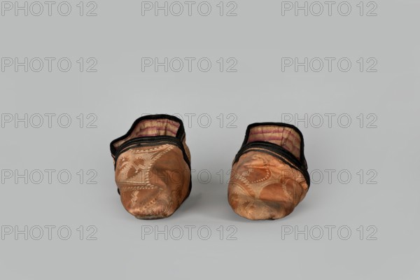 Slippers, origin China, 19th century. Women's footwear: slippers with haberdashery decorations. Lining of the upper and sole made of pink striped canvas; illegible rectangular stamp on the lining. Machine sewing supplemented by hand. Leather sole with a semicircular under-heel piece, tapering sharply to a pointed toe; second layer of leather under the heel nailed with metal nails. Height 5.3 cm, length 17 cm., A pair of soft slippers placed side by side facing forward on a neutral light gray background, each slipper showing a worn orange-brown fabric exterior with decorative stitched patterns, black trim around the opening, and an inner lining with faded pink and beige stripes; the slippers appear slightly misshapen and creased with visible areas of darker discoloration on the toes, and the overall color palette includes orange-brown, black, pink, beige, and gray.