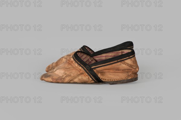 Slippers, origin unknown, haberdashery decorations; women's footwear with lining of the upper and sole made of pink striped canvas. Illegible rectangular stamp on the lining. Machine sewing supplemented by hand. Leather sole, semicircular under the heel, tapering sharply towards the toe to form a point. Under the heel a second layer of leather is nailed with metal nails. 19th century, China. Height 5.3 cm, length 17 cm., A pair of worn slip-on shoes made of tan leather with darker brown scuffing and decorative stitched patterns, trimmed with black fabric around the openings and edges, showing a striped textile lining in shades of red and beige, with flattened soles that are darker brown.