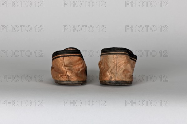 Slippers, origin unknown, women's slippers, haberdashery decorations; lining of the upper and sole made of pink striped canvas with an illegible rectangular stamp on the lining. Machine sewing supplemented by hand. Leather sole, semicircular under the heel, tapering sharply toward a pointed toe. Under the heel a second leather layer is nailed with metal nails. 19th century, China; footwear, clothing; height 5.3 cm, length 17 cm., A pair of worn slip-on shoes viewed from the back sitting on a flat surface against a neutral backdrop; the shoes are primarily tan with darker brown and black accents along the top edges and soles, showing creases and textured patterns on the material, with scuffed and slightly dirty areas on the soles and lower sides, and the background and surface appear in shades of light gray.