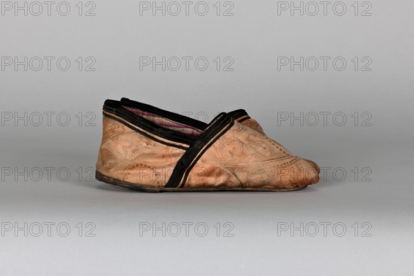 Slippers, origin unknown, 19th century, China. Women's footwear: slippers with haberdashery decorations. Lining of the upper and sole made of pink striped canvas. Illegible rectangular stamp on the lining. Machine sewing supplemented by hand. Leather sole with a semicircular piece under the heel, tapering sharply toward a pointed toe; a second layer of leather under the heel nailed with metal nails. Height 5.3 cm, length 17 cm., A single pair of low, slip-on shoes sits on a smooth, neutral gray surface against a gray backdrop; the shoes are a warm tan color with subtle patterned stitching and worn areas, trimmed with black edging and featuring an interior lining that appears a muted pink or mauve; the soles are darker, closer to black or dark brown, and the shoes show creases and slight scuffing consistent with use.