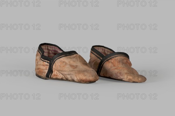 Slippers, origin unknown, 19th century, China. Women's footwear: slippers with haberdashery decorations. Lining of the upper and sole made of pink striped canvas. Illegible rectangular stamp on the lining. Machine sewing supplemented by hand. Leather sole, semicircular under the heel, tapering sharply to a pointed toe. Under the heel a second layer of leather is nailed with metal nails. Height 5.3 cm, length 17 cm., A pair of soft, worn shoes sit on a plain surface, showing a faded peachy-pink fabric with visible wear and discoloration, dark edging around the openings, and a striped inner lining in shades of pink and purple; the soles appear darker and scuffed, and the overall colors visible include peachy-pink, black, pink, purple, and grey from the background.