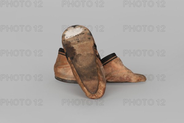 Slippers, origin unknown, haberdashery decorations; women's footwear, slippers. Lining of the upper and sole made of pink striped canvas. Illegible rectangular stamp on the lining. Machine sewing, supplemented by hand. Leather sole, semicircular under the heel, tapering sharply towards the toe to form a point. Under the heel, a second layer of leather is nailed with metal nails. 19th century, China. Height 5.3 cm, length 17 cm., A pair of worn slip-on shoes made of tan leather with darker brown scuffing and stains, one shoe positioned sole-up showing a dark brown worn sole with metal rivets around the heel, both shoes having black trim near the openings and areas of creasing and discoloration, set against a plain light gray background.
