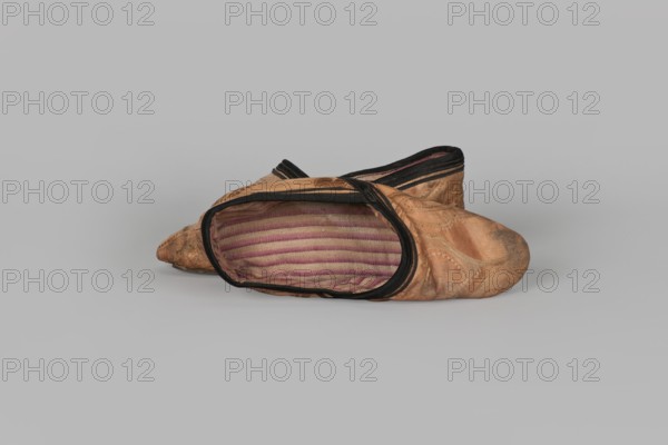 Slippers, origin unknown, 19th century, China. Women's footwear: slippers with haberdashery decorations. Lining of the upper and sole made of pink striped canvas with an illegible rectangular stamp on the lining. Machine sewing supplemented by hand. Leather sole, semicircular under the heel, tapering sharply to a pointed toe. Under the heel a second layer of leather is nailed with metal nails. Height 5.3 cm, length 17 cm., A worn pair of slip-on shoes resting on a flat surface, one shoe positioned slightly in front of the other with the opening of the front shoe facing the viewer to reveal a striped interior lining; the exterior shows a textured, patterned fabric with visible creases and scuffs, and the shoe edges are trimmed with a dark band. The visible colors include tan, brown, black, pink, and beige.