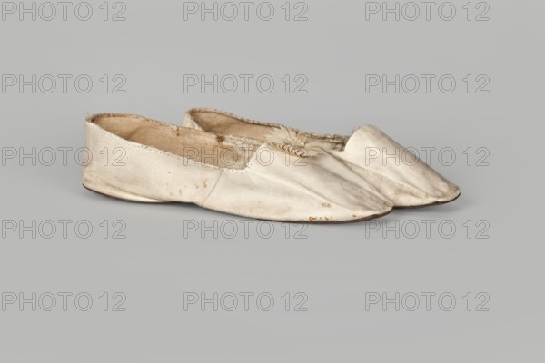Women’s slippers, maker: Johann Dyck Vilnius; shoemaker’s workshop; ca. 1800–ca. 1850. Flat women’s slippers made of white full-grain leather with flat-cut toes, decorated with a rosette of white grosgrain ribbon one rosette preserved. Symmetrical leather soles. White canvas lining and endpaper. A printed company label is glued onto the endpaper of one slipper: “Johann / Dyck / …auf der / Schloss-strase / nr … in / VILNIUS.” 1st half of the 19th century 19th century, place of origin: Vilnius. Footwear, clothing; height 6 cm, length 24 cm., A pair of flat, slip-on shoes made of a light-colored material, shown side by side on a neutral background; the shoes are pale cream to off-white with visible wear and small dark spots, fraying at the inner edges, and slightly darker, beige interior linings; one shoe has a decorative element near the opening made of layered, petal-like pieces in similar pale tones, and the soles appear thin with a slightly darker brown edge.