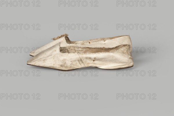 Women’s slippers. Maker: Johann Dyck, shoemaker’s workshop in Vilnius ca. 1800–ca. 1850. Flat women’s slippers made of white full-grain leather with flat-cut toes, decorated with a rosette of white grosgrain ribbon one rosette preserved. Symmetrical leather soles, white canvas lining and endpaper. A printed company label is glued onto the endpaper of one slipper: “Johann / Dyck / …auf der / Schloss-strase / nr … in / VILNIUS.” Date: 1st half of the 19th century 19th century. Type/place of origin: Vilnius. Category: footwear, clothing; women’s footwear/slippers. Dimensions: height 6 cm; length 24 cm., A pair of worn slip-on shoes made of a light material, shown from the side and overlapping, with pointed toes and low flat soles; the shoes display visible creases, scuffs, and frayed edges, especially around the openings, and the interior lining is partly exposed; the dominant colors are off-white and beige with areas of darker dirt and brownish discoloration, and the soles appear darker ...