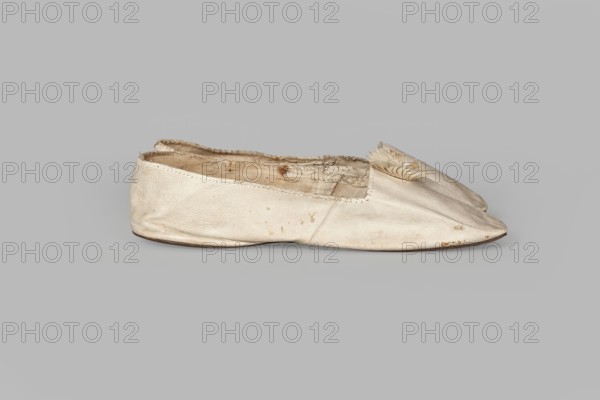 Women’s slippers. Maker: Johann Dyck, shoemaker’s workshop, Vilnius; ca. 1800–ca. 1850. Flat women’s slippers made of white full-grain leather with flat-cut toes, decorated with a rosette of white grosgrain ribbon one rosette preserved. Symmetrical leather soles. White canvas lining and endpaper. A printed company label is glued onto the endpaper of one slipper: “Johann / Dyck / …auf der / Schloss-strase / nr … in / VILNIUS.” 1st half of the 19th century. Materials: leather, grosgrain ribbon, canvas. Dimensions: height 6 cm, length 24 cm., A single flat shoe is shown from a side view resting on a plain gray background, made of a light cream or off-white material with visible wear and slight discoloration; the upper has a soft, wrinkled texture and a few small darker spots, and the interior lining appears frayed and stained with brownish marks; the toe area is pointed and decorated with a small folded fabric detail in a similar cream color, and the sole edge is a darker brown.
