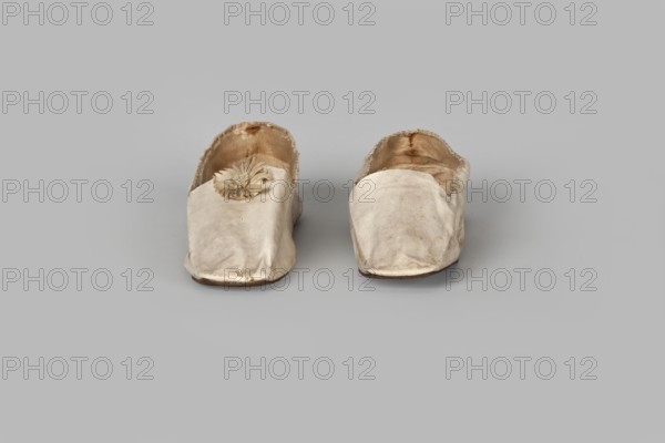 Women’s slippers by Johann Dyck, shoemaker’s workshop, Vilnius, ca. 1800–ca. 1850. Flat women’s slippers made of white full-grain leather with flat-cut toes, decorated with a rosette of white grosgrain ribbon one rosette preserved. Symmetrical leather soles. White canvas lining and endpaper. A printed company label is glued onto the endpaper of one slipper: “Johann / Dyck / …auf der / Schloss-strase / nr … in / VILNIUS.” 1st half of the 19th century. Footwear, clothing; height 6 cm, length 24 cm., A pair of slip-on shoes sitting side by side on a plain background, appearing worn and made of a pale fabric with visible creases and small stains; the left shoe has a decorative rosette on its top, while the interiors show frayed edges and darker discoloration; colors visible include off-white, beige, brown, and gray in the background.