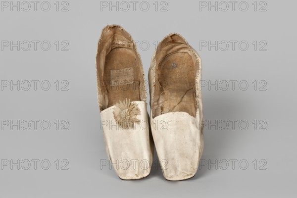 Women’s slippers, maker: Johann Dyck Vilnius; shoemaker’s workshop; ca. 1800–ca. 1850. Flat women’s slippers made of white full-grain leather, 1st half of the 19th century. Flat-cut toes, decorated with a rosette of white grosgrain ribbon one rosette preserved. Symmetrical leather soles. White canvas lining and endpaper. A printed company label is glued onto the endpaper of one slipper: “Johann / Dyck / …auf der / Schloss-strase / nr … in / VILNIUS.” Height 6 cm; length 24 cm. Type/place of origin: Vilnius. Materials/subjects: haberdashery decorations, company labels, leather goods, slippers, footwear, women’s footwear, women’s slippers, fashion, clothing accessories., A pair of worn slip-on shoes shown from above on a plain background, the shoes are primarily off-white with visible areas of beige and brown on the interiors, the outer material appears smooth with some scuffs and slight discoloration, one shoe has a decorative rosette made of the same off-white material near the opening...