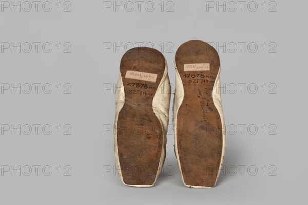 Women’s slippers, maker: Johann Dyck Vilnius; shoemaker’s workshop; ca. 1800–ca. 1850. Flat women’s slippers made of white full-grain leather. Flat-cut toes, decorated with a rosette of white grosgrain ribbon—one rosette preserved. Symmetrical leather soles. White canvas lining and endpaper. A printed company label is glued onto the endpaper of one slipper: “Johann / Dyck / …auf der / Schloss-strase / nr … in / VILNIUS.” 1st half of the 19th century 19th century, Vilnius. Footwear, clothing; height 6 cm, length 24 cm., A pair of shoes shown from the soles, positioned side by side on a plain background; the soles are brown and worn with visible scuffs and darker spots, and the shoe uppers visible around the edges are an off-white or cream color; the background is a uniform light gray.