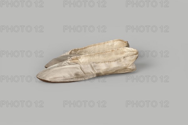 Flat women's slippers of white satin with top edge finished with ribbon. Flat-cut toes, symmetrical leather soles. Endpaper and back lining of white leather; front lining of canvas. On the endpaper of both slippers there is an octagonal, oblong company label: Jan Kólazenski / in Kalisz / at Market Square No. 18. Maker: Jan Kólazenski Kalisz; shoemaker's workshop, ca. 1830–1850. Type: women's footwear; height 6 cm, length 23.7 cm., A pair of soft, worn slip-on shoes made of light-colored fabric sit side by side on a smooth gray background; the shoes show creases and areas of darker discoloration, with visible stitching along the edges and slightly uneven, flattened soles. The visible colors are shades of off-white, beige, cream, light tan, and gray.