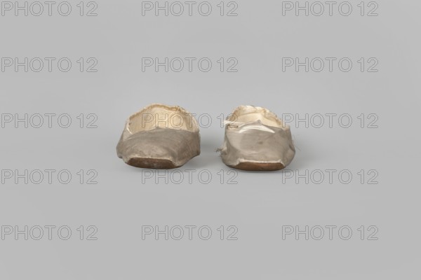 Flat women's slippers of white satin, top edge finished with ribbon. Flat-cut toes, symmetrical leather soles. Endpaper and back lining of white leather, front lining of canvas. On the endpaper of both slippers there is an octagonal, oblong company label: Jan Kólazenski / in Kalisz / at Market Square No. 18. Maker: Jan Kólazenski, Kalisz; shoemaker's workshop; ca. 1830–ca. 1850. Function: women's footwear, clothing accessory. Dimensions: height 6 cm; length 23.7 cm., A pair of worn slip-on shoes sit side by side on a flat surface, each showing a soft, creased upper with frayed edges and a visible inner lining; the shoes are primarily light beige to off-white with darker beige and brown areas on the soles and toe regions, and the background is a uniform light gray.