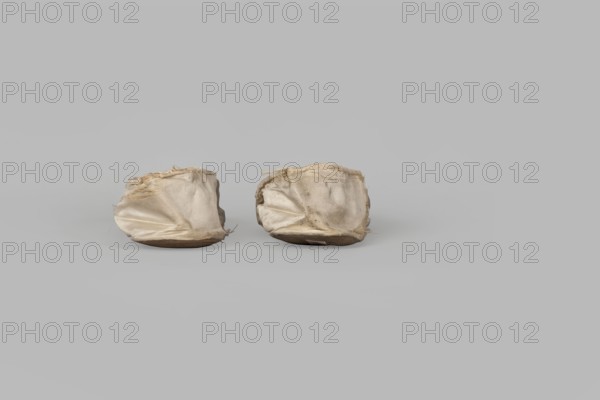 Flat women's slippers of white satin, top edge finished with ribbon. Flat-cut toes, symmetrical leather soles. Endpaper and back lining of white leather, front lining of canvas. On the endpaper of both slippers there is an octagonal, oblong company label: Jan Kólazenski / in Kalisz / at Market Square No. 18. Maker: Jan Kólazenski, Kalisz; shoemaker's workshop; ca. 1830–1850. Women's footwear, clothing accessory; height 6 cm, length 23.7 cm., A pair of small fabric items sit side by side on a flat surface, each appearing soft and slightly collapsed with wrinkled, worn cloth that has frayed edges; the fabric shows shades of off-white, beige, and light tan with some darker smudges, and each item rests on a low, rounded base that is brown; the background and surface are a uniform light gray.