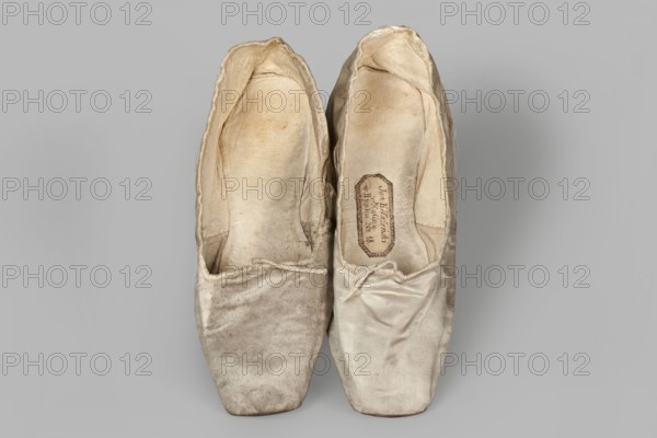 Flat women's slippers of white satin, top edge finished with ribbon. Flat-cut toes, symmetrical leather soles. Endpaper and back lining of white leather, front lining of canvas. On the endpaper of both slippers there is an octagonal, oblong company label: Jan Kólazenski / in Kalisz / at Market Square No. 18. Maker: Jan Kólazenski Kalisz; shoemaker's workshop; ca. 1830–ca. 1850. Materials: satin, leather, canvas. Type: women's footwear, slippers. Dimensions: height 6 cm, length 23.7 cm., A pair of worn ballet slippers photographed from above, showing the insides and toes; the slippers are made of a light, satin-like material with a slightly shiny texture and visible creases and folds; the left slipper appears more discolored and dirty than the right, with darker smudging and staining on the toe and sides, while the right slipper is cleaner with lighter soiling; the inner linings look soft and aged with uneven pigmentation and a few spots; a thin drawstring is visible at the vamp of each...