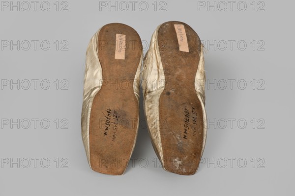 Flat women's slippers of white satin, top edge finished with ribbon. Flat-cut toes, symmetrical leather soles. Endpaper and back lining of white leather, front lining of canvas. On the endpaper of both slippers there is an octagonal, oblong company label: Jan Kólazenski / in Kalisz / at Market Square No. 18. Maker: Jan Kólazenski Kalisz; shoemaker's workshop; ca. 1830–ca. 1850. 1830–1850. Place of origin: Kalisz. Materials: satin, leather, canvas. Function: women's footwear slippers. Dimensions: height 6 cm, length 23.7 cm., A pair of worn shoes shown from the soles, laid side by side on a plain light gray background; the shoe uppers are a pale ivory or off-white fabric with some fraying at the edges, and the soles are a brown leather with darker scuff marks and patches of discoloration; there are a few small dark smudges and worn areas on the soles and slightly uneven edges where the fabric meets the sole.