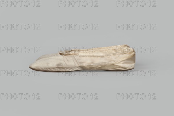 Flat women's slippers of white satin with top edge finished with ribbon. Flat-cut toes, symmetrical leather soles. Endpaper and back lining of white leather, front lining of canvas. On the endpaper of both slippers there is an octagonal, oblong company label: Jan Kólazenski / in Kalisz / at Market Square No. 18. Maker: Jan Kólazenski, shoemaker's workshop, Kalisz; ca. 1830–1850. Function: women's footwear; materials: satin, leather, canvas. Dimensions: height 6 cm, length 23.7 cm., A single worn shoe-like textile object rests on a flat surface against a plain background, showing a soft, slightly wrinkled fabric exterior in shades of off-white and beige with areas of darker beige and faint grayish smudging; the shape is elongated and flattened with a pointed toe and a low, rounded heel, seams and stitching are visible along the top edge and side, and there are loose threads and frayed fabric near the opening and along some seams.
