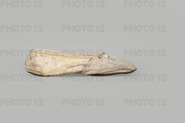 Flat women's slippers of white satin, top edge finished with ribbon. Flat-cut toes, symmetrical leather soles. Endpaper and back lining of white leather, front lining of canvas. On the endpaper of both slippers there is an octagonal, oblong company label: Jan Kólazenski / in Kalisz / at Market Square No. 18. Made by Jan Kólazenski, Kalisz; shoemaker's workshop, ca. 1830–1850. Women's footwear, slippers; height 6 cm, length 23.7 cm., A worn fabric slipper sits sideways on a plain background, showing a rounded toe and a low, flat sole; the fabric appears creased and frayed with visible stitching along the top edge and a small tied bow near the opening. The slipper's surface shows uneven discoloration and spots, with areas of dirt and wear, and some loose threads along the seams and lower edge. The visible colors are off-white, beige, light brown, and gray, with a darker brown sole.