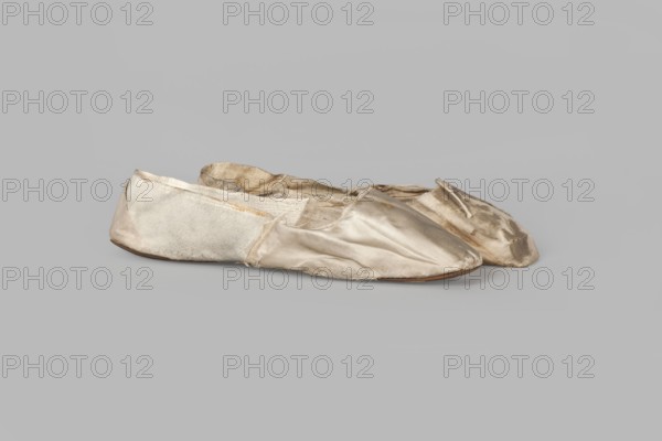 Flat women's slippers of white satin. Flat-cut toe, symmetrical thin leather soles. Endpaper and back of the lining are white glacé leather; front lining is white canvas. On the endpaper an octagonal elongated company label printed in green reads: Józef Marczewski / in Warsaw / at Dluga Street / No. 543. Maker: Józef Marczewski Warsaw, shoemaker's workshop, ca. 1830–ca.1840. Date: 1836. Place of origin: Warsaw. Material/technique: satin, leather, canvas. Function: women's footwear slippers. Dimensions: height 5.2 cm; length 23.5 cm., A pair of worn soft shoes made of a shiny fabric in shades of off-white, beige, and pale cream sit on a flat neutral gray surface; the shoes show fraying edges, creases, and areas of discoloration in light brown and tan, with a slightly darker brown sole visible beneath, and the fabric texture varies between smooth shiny panels and more matte, slightly fuzzy sections.