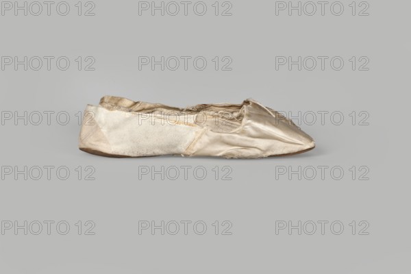Flat women's slippers of white satin, ca. 1836. Flat-cut toe, symmetrical thin leather soles. Endpaper and back of lining made of white glacé leather; front of lining white canvas. Endpaper bears an octagonal elongated company label printed in green with inscription: Józef Marczewski / in Warsaw / at Dluga Street / No. 543. Height 5.2 cm; length 23.5 cm., A single worn ballet slipper rests on a neutral grey background, showing a satin-like upper with visible creases and fraying along the edges, a softer fabric panel along the side, and a thin darker sole; the slipper displays shades of beige, cream, off-white, light tan, and a hint of brown on the sole, with subtle sheen on the satin areas and a slightly matte texture on the fabric panel.