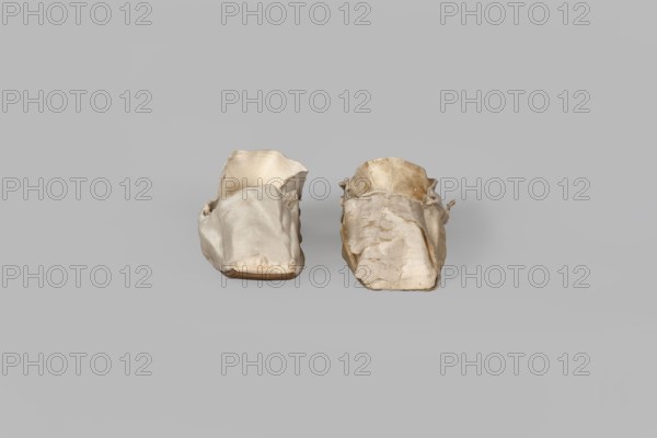 Women's slippers. Maker: Józef Marczewski, shoemaker's workshop, ca. 1830–ca. 1840. Flat women's slippers of white satin with flat-cut toe and symmetrical thin-leather soles. Lining: endpaper and back in white glacé leather, front in white canvas. Endpaper bears an octagonal elongated company label printed in green: Józef Marczewski / in Warsaw / at Dluga Street / No. 543. Date: 1836, place of origin: Warsaw. Materials: satin, leather, canvas. Dimensions: height 5.2 cm, length 23.5 cm. Function: footwear, women's fashion accessory., A pair of small, worn fabric shoe covers sit side by side on a plain light gray background; the covers are off-white to beige in color with areas of darker staining and fraying, uneven edges, and creased, wrinkled surfaces, and the inner linings visible in places show slightly different lighter beige tones, while the soles appear to be a muted tan.