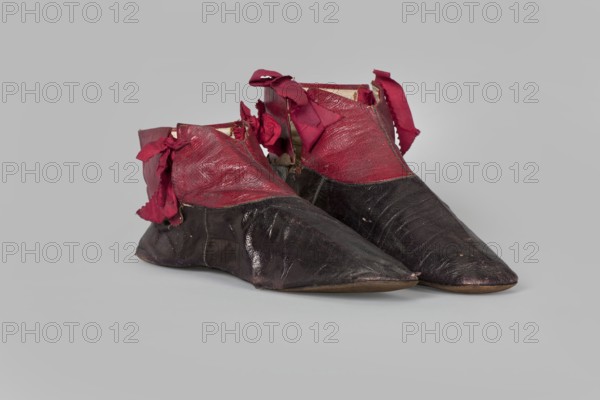 Women's ankle boots made of graphite full-grain leather with red leather uppers tied on the outside and inside with a red ribbon. Thin, symmetrical leather soles with bevelled toes and no heels. Canvas lining, white leather endpapers. On the endpaper an oval company label reads: Berlin / 23 Jäger str / Fabrique de chassures / pour les Dames / Berlin / 23 Jäger str / Damen / Schuh-fabrik von / CP Schröder. The upper is machine-sewn; the lining is hand-sewn. Ca. 1850, Berlin. Footwear; clothing. Dimensions: height 10 cm, width 5.5 cm, length 25.5 cm., A pair of pointed shoes with a glossy dark lower portion and a textured upper portion in a deep red, each shoe showing frayed red fabric ties and rough edges, set against a pale gray background with soft shadows beneath.
