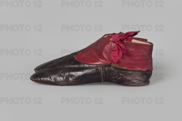Women's ankle boots made of graphite full-grain leather with red leather uppers tied on the outside and inside with a red ribbon. Thin, symmetrical leather soles with bevelled toes and no heels. Canvas lining, white leather endpapers. On the endpaper there is an oval company label: Berlin / 23 Jäger str / Fabrique de chassures / pour les Dames / Berlin / 23 Jäger str / Damen / Schuh-fabrik von / CP Schröder. The upper part is machine-sewn; the lining is hand-sewn. Ca. 1850. Footwear, women's ankle boots; height 10 cm, width 5.5 cm, length 25.5 cm., A pair of low-cut shoes placed side by side on a pale gray background, made of glossy dark brown leather on the lower portion and a textured red leather on the upper portion, with a frayed red fabric tied in a bow near the ankle area; the dark brown leather shows creases and wear while the red leather appears smoother with some scuffs, and a strip of worn tan material is visible along the seam between the two leathers.