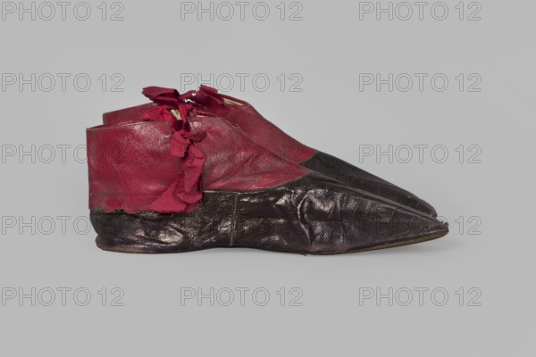 Women's ankle boots made of graphite full-grain leather with red leather uppers tied on the outside and inside with a red ribbon. Thin, symmetrical leather soles with bevelled toes and no heels. Canvas lining, white leather endpapers. On the endpaper there is an oval company label: Berlin / 23 Jäger str / Fabrique de chassures / pour les Dames / Berlin / 23 Jäger str / Damen / Schuh-fabrik von / CP Schröder. The upper part is machine-sewn; the lining is hand-sewn. Circa 1850, Berlin. Footwear; clothing. Dimensions: height 10 cm, width 5.5 cm, length 25.5 cm., A pair of pointed shoes photographed from the side resting on a pale gray background, showing uppers made of a deep red material with torn red fabric ties and lower sections made of a dark glossy material that appears black with a hint of brown; the shoes show creases and worn texture, with visible seams and slightly raised soles, and the image includes colors red, black, brown, and gray.