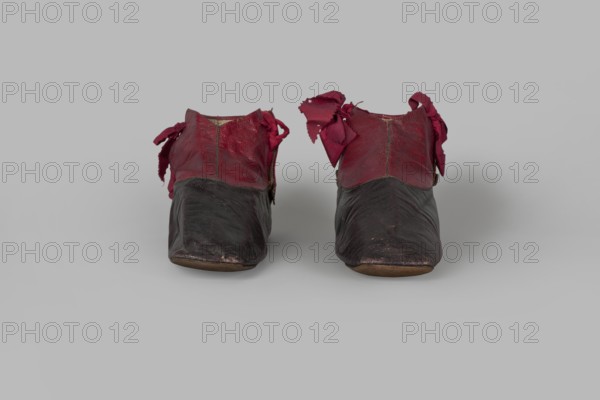 Women's ankle boots made of graphite full-grain leather with red leather uppers tied on the outside and inside with a red ribbon. Thin, symmetrical leather soles with bevelled toes and no heels. Canvas lining, white leather endpapers. On the endpaper there is an oval company label: Berlin / 23 Jäger str / Fabrique de chassures / pour les Dames / Berlin / 23 Jäger str / Damen / Schuh-fabrik von / CP Schröder. The upper part is machine-sewn, the lining is hand-sewn. Circa 1850, Berlin. Footwear, women's ankle boots; height 10 cm, width 5.5 cm, length 25.5 cm., A pair of small worn shoes displayed side by side on a plain light gray surface, each shoe showing a dark brown lower portion with visible creasing and scuffed areas, and a deep red upper section with frayed fabric bows at the sides; the shoes have rounded toes and low profiles, and the visible colors include dark brown, deep red, light brown on the soles, and the surrounding light gray background.