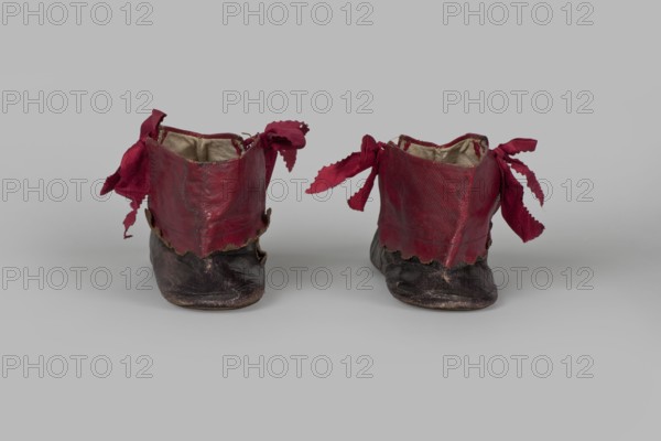 Women's ankle boots made of graphite full-grain leather with red leather uppers tied on the outside and inside with a red ribbon. Thin, symmetrical leather soles with beveled toes and no heels. Canvas lining, white leather endpapers. On the endpaper there is an oval company label: Berlin / 23 Jäger str / Fabrique de chassures / pour les Dames / Berlin / 23 Jäger str / Damen / Schuh-fabrik von / CP Schröder. The upper part is machine-sewn, the lining is hand-sewn. Ca. 1850, Berlin. Footwear, clothing; height 10 cm, width 5.5 cm, length 25.5 cm., A pair of small worn shoes sit side by side on a neutral gray surface against a gray background, each shoe featuring a dark lower portion in blackish brown and an upper section in deep red leather with scalloped edges; loose red fabric ties extend from the sides of both shoes with frayed ends, and the interiors show a lighter beige lining.