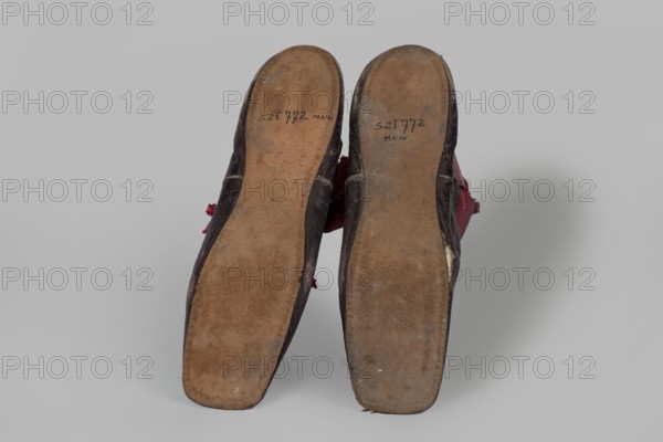 Women's ankle boots made of graphite full-grain leather with red leather uppers tied on the outside and inside with a red ribbon. Thin, symmetrical leather soles with bevelled toes and no heels. Canvas lining, white leather endpapers. On the endpaper there is an oval company label: Berlin / 23 Jäger str / Fabrique de chassures / pour les Dames / Berlin / 23 Jäger str / Damen / Schuh-fabrik von / CP Schröder. The upper part is machine-sewn, the lining is hand-sewn. Circa 1850, Berlin. Footwear; women's ankle boots. Dimensions: height 10 cm, width 5.5 cm, length 25.5 cm., A pair of worn shoes shown from the soles, positioned side by side against a light gray background; the soles are brown and scuffed, the shoe uppers visible at the edges are dark brown with hints of red fabric peeking out, and there are areas of lighter wear and discoloration on the soles and edges.