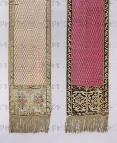 Kontusz belt, ; carp scale ornament; flower bushes ornament; ribbon ornament; bipartite heads; solid, reversible Polish kontusz belt; men's clothing accessory; bipartite heads with a motif of stylized flower bushes surrounded by a wavy ribbon. The belt is woven in a carp scale pattern. Sewn-on gold fringe. Colors: brown, gold, red, green. 18th century, Lyon, France; national costume accessory. Length 353 cm, width 30 cm., Two long narrow fabric panels hang side by side against a light background; the left panel is primarily cream with a small repeating scaled pattern and a decorative floral border along its edges and bottom, and the right panel is primarily deep pink with a similar scaled pattern framed by a dark floral border. Both panels have ornate rectangular sections near the bottom with stylized floral motifs in colors including pink, blue, green, cream, gold, and brown, and both end in long gold-colored fringe. The left panel shows an overall pale cream and gold appearance with ...