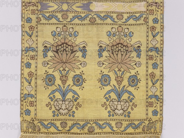 Kontusz sash by Paschalis Jakubowicz Manufactory Warsaw; belt manufacturer; 1788–1794. Maker: Jakubowicz, Jakub Paschalis ?-1816/1817. Polish kontusz sash four-sided sash for national/mens' costume, Lipków Masovian Voivodeship origin, dated 1791–1794. Description: bipartite heads with the pattern of elongated flower bouquets; transverse stripes with alternating floral and geometric ornamentation; flower bushes ornament. Sewn-on silk fringe, beige. Colors: straw, pink, blue, light green. Materials: silk. Function: clothing/fashion accessory. Dimensions: length 272 cm, width 36 cm., A rectangular textile with a pale yellow background decorated with two mirrored stylized floral compositions in muted brown, blue, cream, and tan tones, each composition featuring a central large leaf-like flower with radiating petals, smaller round flowers, drooping blue leaves, and clustered berry-like shapes; a narrow floral border runs along the left and right edges with alternating small flower motifs in...
